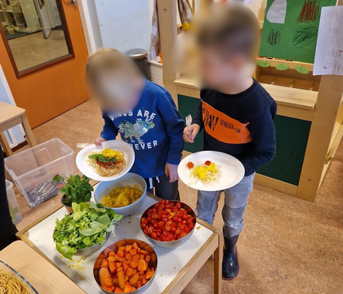 Buffet in de klas met verschillende kleuren groenten en soorten pasta waarvan de kinderen opscheppen.